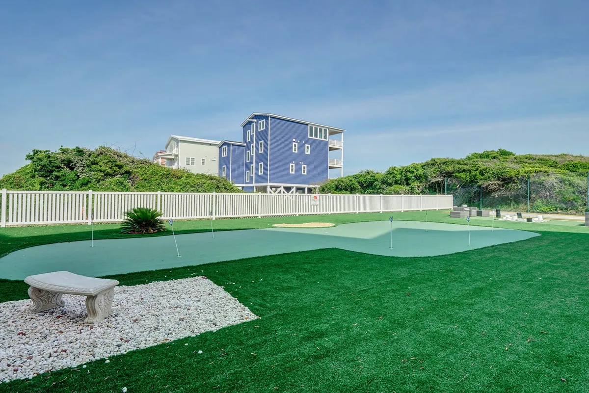 An inviting view of the putting green adjacent to a blue house, with a stone bench and manicured landscaping highlighting a leisurely and fun outdoor atmosphere ideal for golf practice and social interaction.