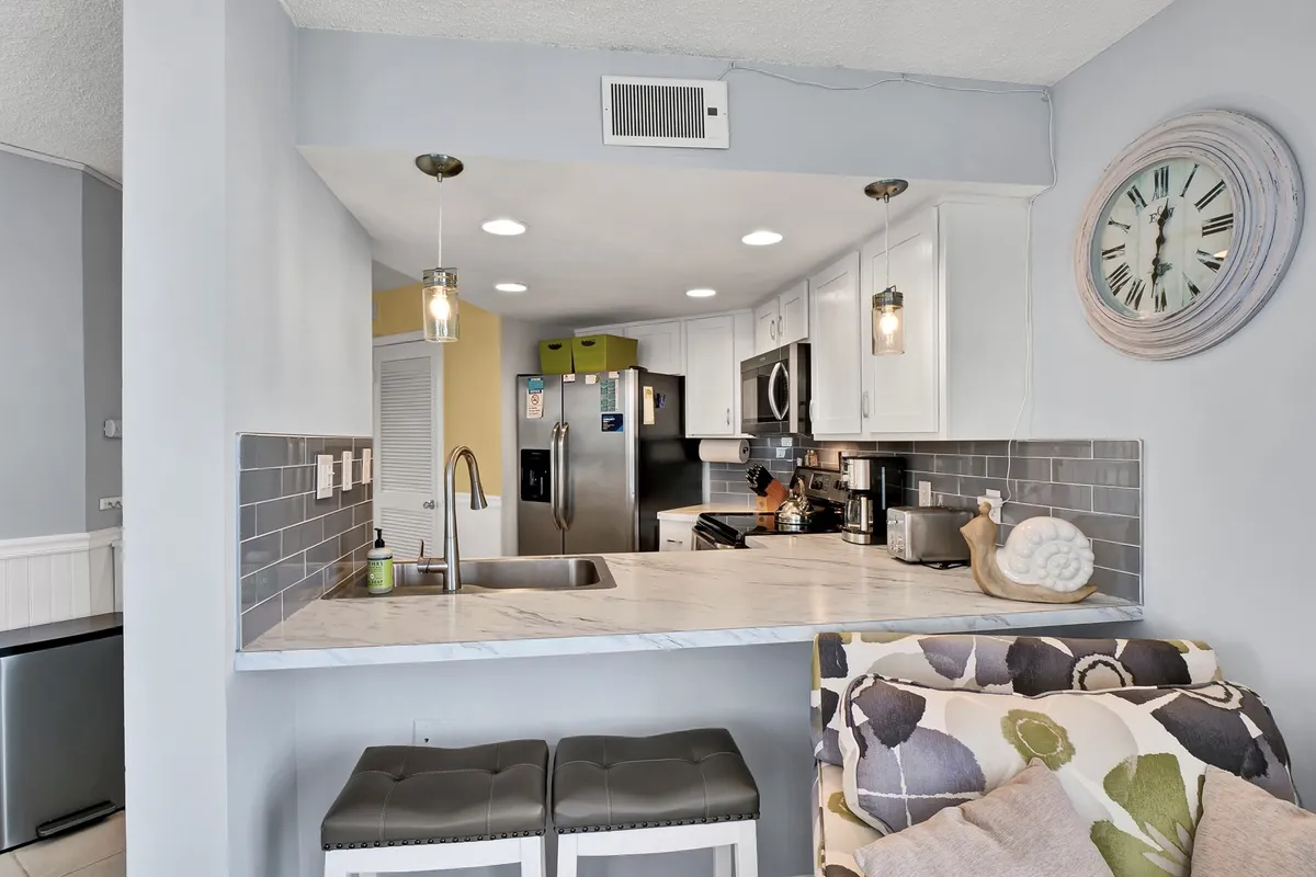 A bright, welcoming kitchen featuring a central island with bar stools, gleaming stainless steel appliances, marble countertops, and a large decorative clock. The space is styled for comfort and functionality, inviting both cooking and casual dining.