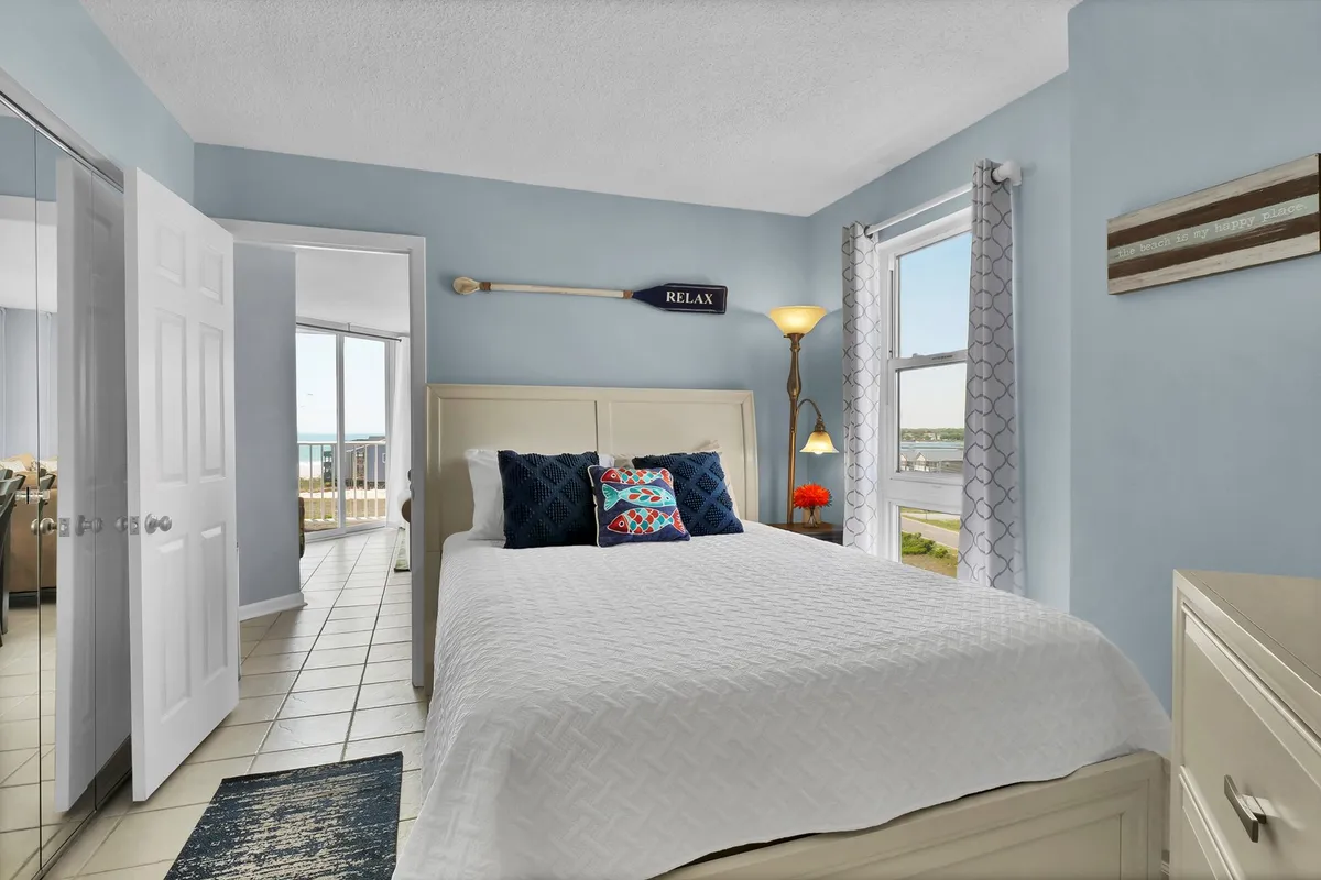 A serene, private master bedroom features a queen bed adorned with decorative pillows and a window inviting in soft light and a view. A lamp and calming 'RELAX' sign enhance the restful, tranquil mood, ideal for a restorative retreat after a long day.