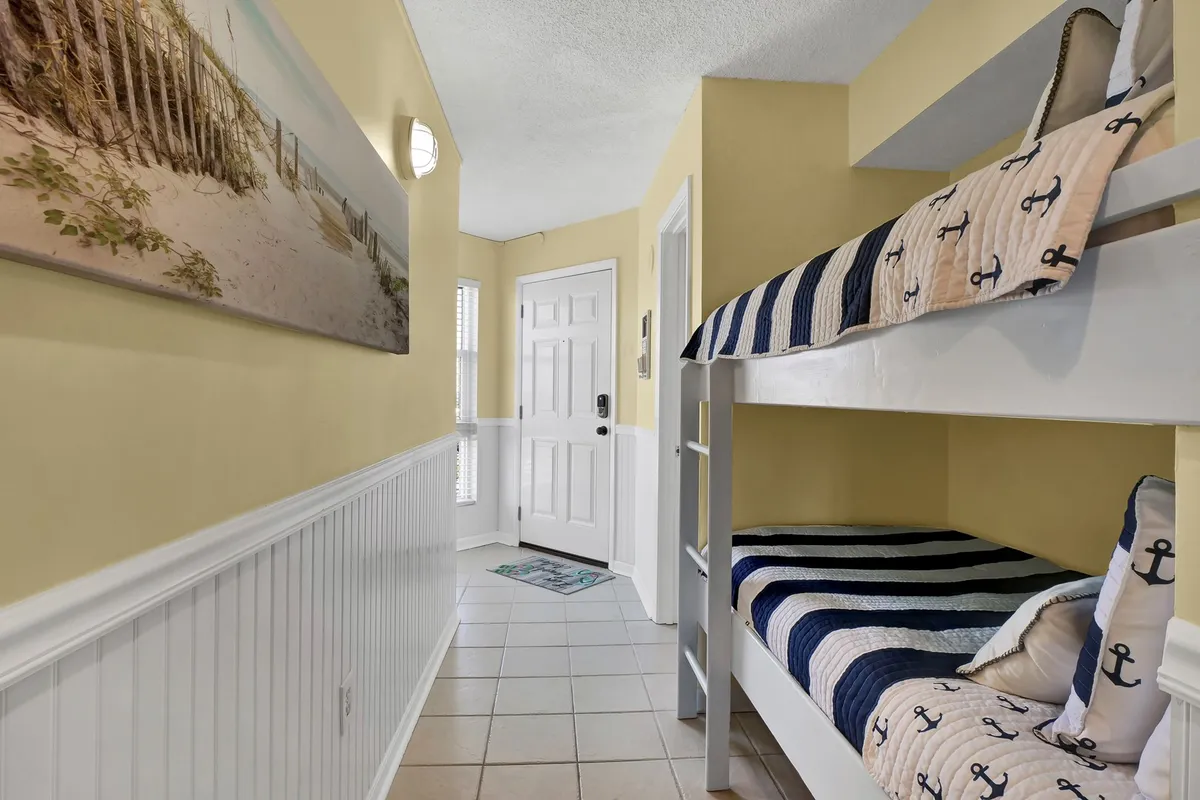This cozy interior scene centers on a set of twin bunk beds, adorned with beach-themed wall art and surrounded by a soft, inviting ambiance. Children are seen playing in the bunk beds, which are nestled in a nook near a doorway, emphasizing a family-friendly, playful environment.