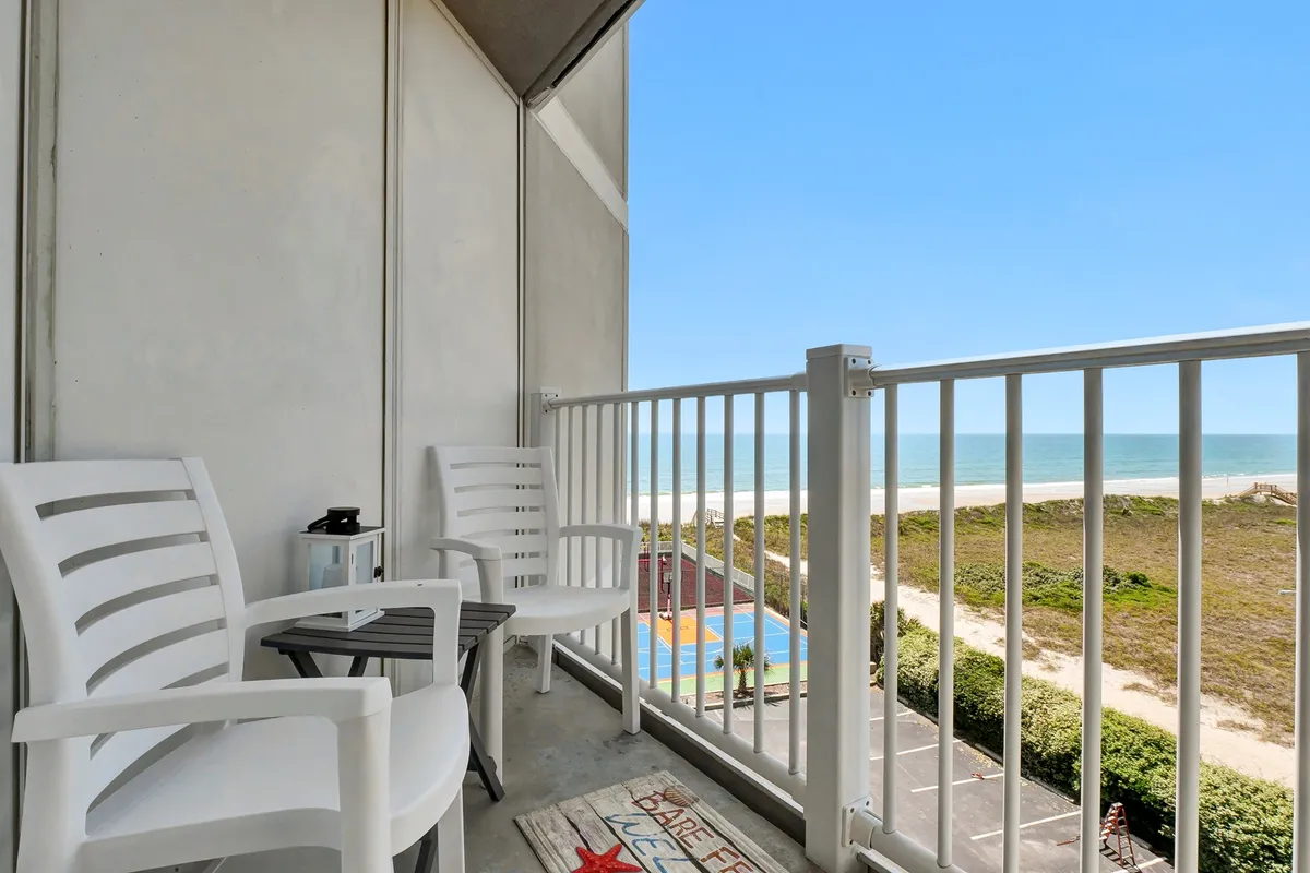 This image captures two balcony chairs facing a stunning, unobstructed ocean view with a colorful beach area below. The serene composition invites the viewer to imagine sipping morning coffee or relaxing together while watching the waves and the sky change colors.