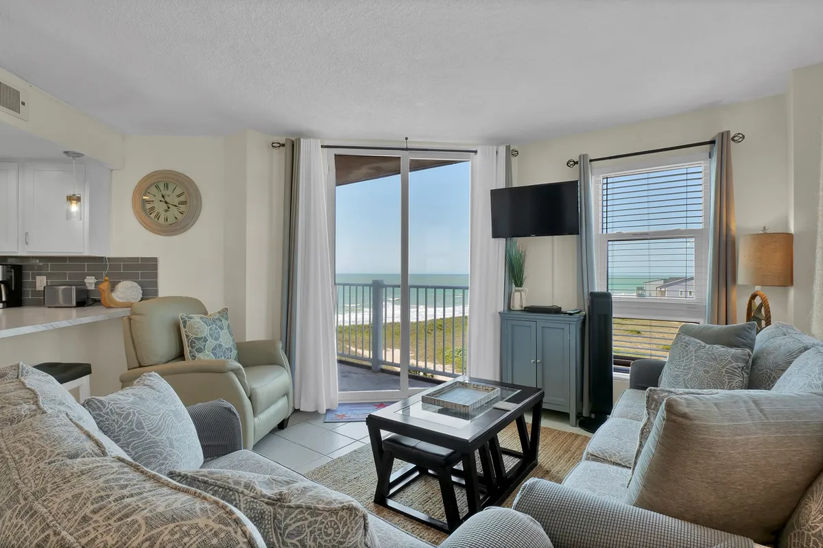 A sunlit living lounge features large windows framing panoramic ocean views. Comfortable seating, including a plush couch and coffee table with tasteful decor, invite guests to relax. The tranquil ambiance is enhanced by natural light streaming in, suggesting perfect moments for savoring coffee or reading by the window.