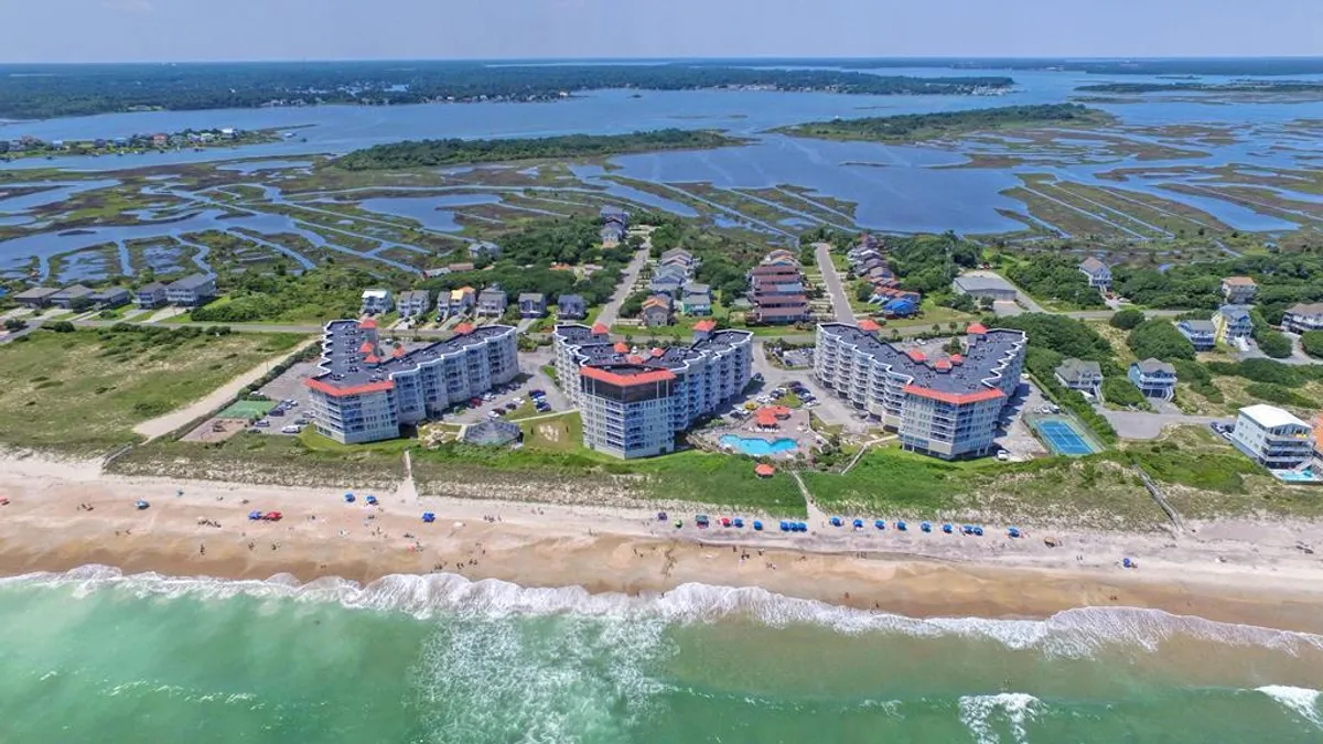 A sweeping exterior view of beachfront buildings with a sparkling swimming pool in the foreground, dotted with beach umbrellas. The scene opens out to a wide, sandy shore and gentle ocean waves, with hints of marshland visible in the background. Families can be seen swimming, sunbathing, and enjoying walks along the beach.
