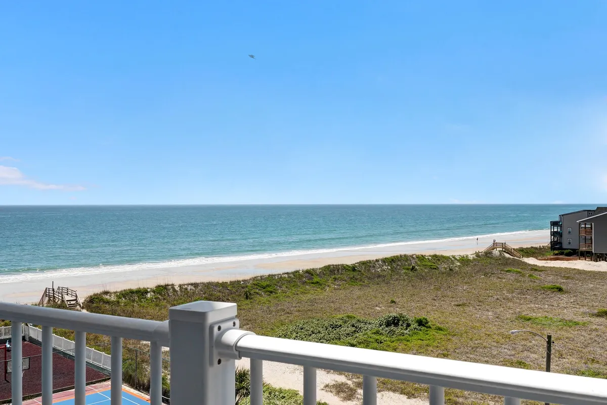 The image captures a serene, sweeping ocean view from a private condo balcony at North Topsail Beach, framed by lush dune vegetation and a sturdy railing. The tranquil scene features the sun rising over the water, pristine sandy shores, and hints of beach activities like sunbathing and strolling, all contributing to a peaceful coastal escape.