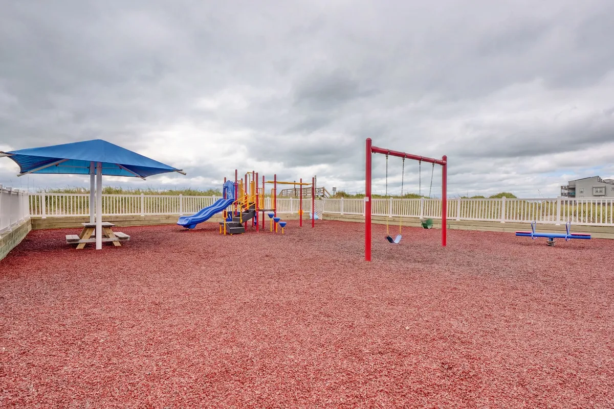 A vibrant playground area set within a secure, fenced section, featuring swings, modern play structures, and a picnic table. The area is filled with children playing and families gathering for picnics, all beneath a sunny sky.
