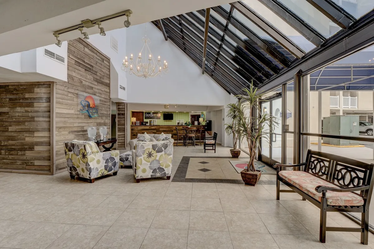 A bright, welcoming interior space featuring a chandelier, a reception desk, comfortable seating, and large windows letting in natural light. The ambiance is both inviting and relaxed, suggesting a resort-like atmosphere for arrivals and casual gatherings.