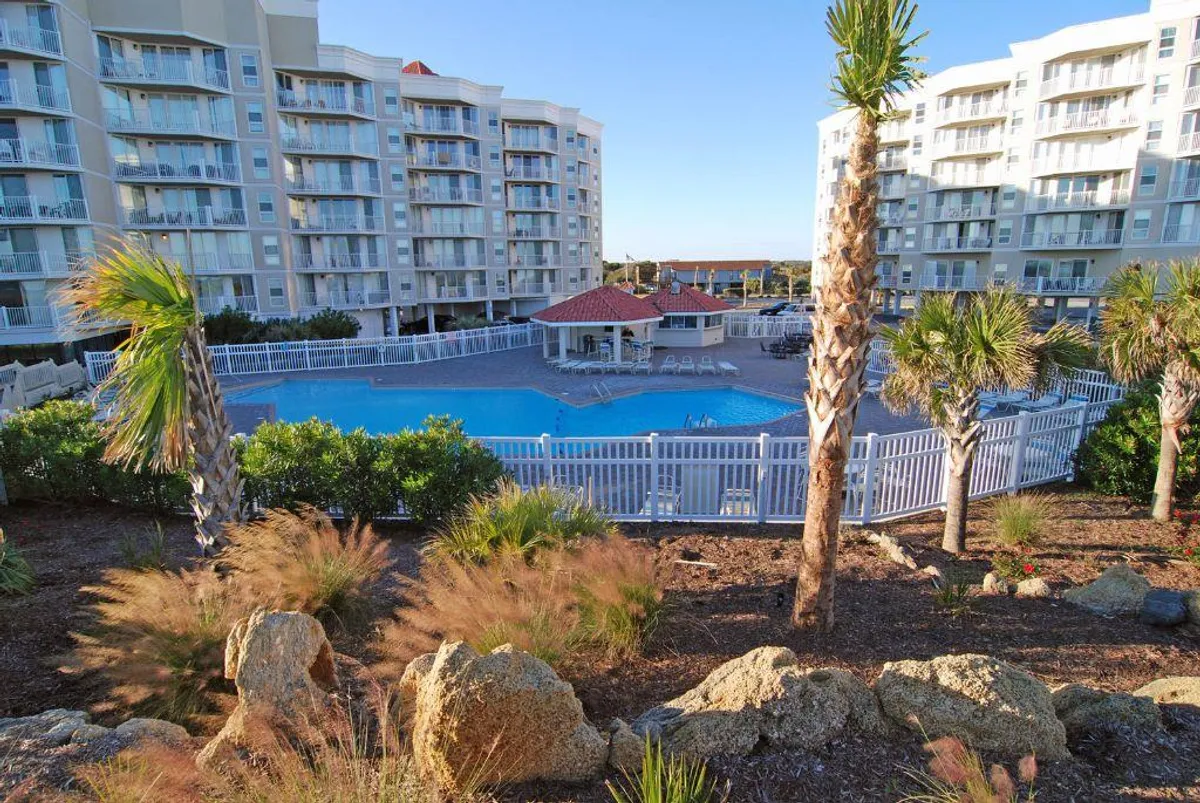 A sunlit scene of the outdoor pool surrounded by palm trees and landscaped gardens, with adjacent buildings in the background. Guests are seen relaxing by the pool and enjoying the tranquil setting.