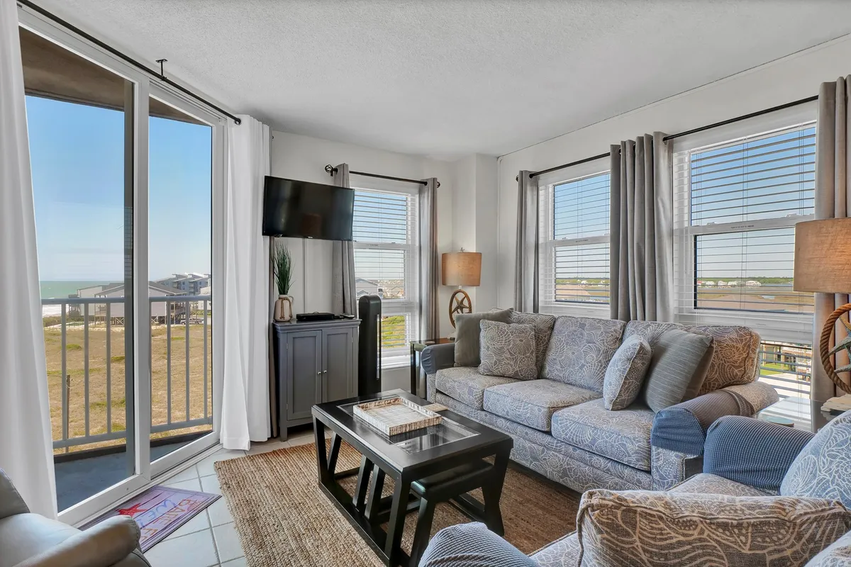 The living area is centered around a sofa and coffee table set in front of wide windows that reveal serene ocean vistas. The presence of a TV hints at cozy movie nights, while the soft morning light suggests an idyllic space for leisurely mornings or gathering with loved ones.